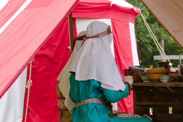 A girl in a rich medieval green dress with her hair dines while sitting at a wooden table in a tent camp. Pottery on the table. Around the red and white tents.