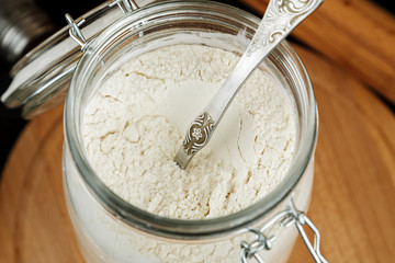 Open glass jar with white wheat flour and metal spoon in it on wooden table among other kitchen stuff