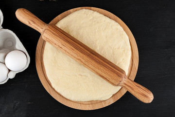 Raw fresh dough on wooden cutting board on black table, pizza cooking process, top view