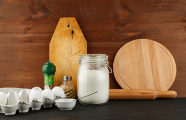 Open glass jar with white wheat flour on black wooden table among other kitchen stuff
