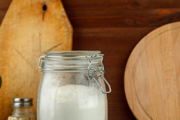 Glass jar with white wheat flour on black wooden table among other kitchen stuff
