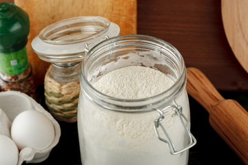 Open glass jar with white wheat flour among other kitchen stuff