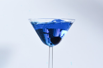 Cocktail with blue liquid in glass. Glass with blue water pouring with liquid with splashes and drops. Martini glass filling with alcohol with splashes on white background. Refreshing drink concept.