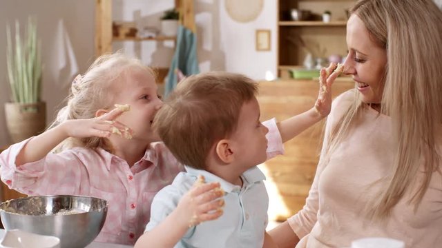 Medium shot of loving young Caucasian mom making dough for baking with her kids, and letting gap-toothed blonde daughter touch her face and nose with her messy hand