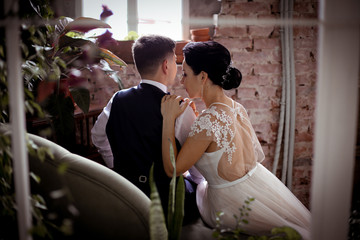 Naklejka premium Groom in vest and beautiful bride in an elegant dress sitting back on the sofa among the houseplants in a stylish interior