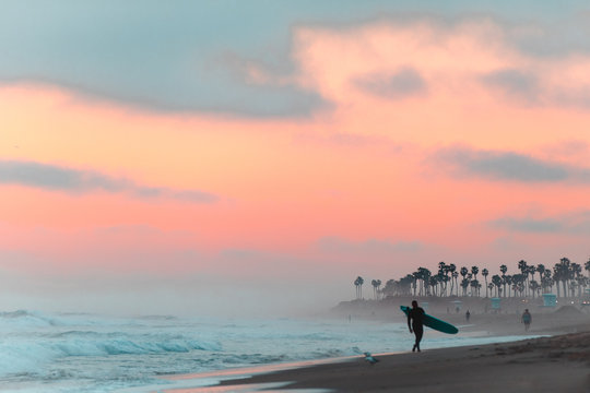 Man walks on beach with surfboard at sunrise