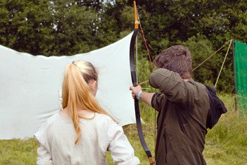 A girl in folk costume is watching a man in modern clothes shooting a backpack from a medieval bow....