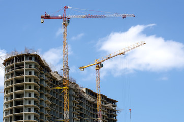 two colored high-altitude cranes on the background of the cloudy sky and the building under construction.