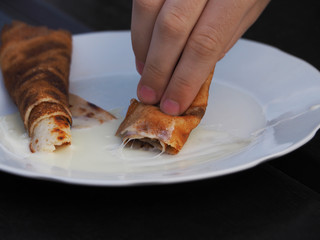 The child's hand holds the pancake wrapped in a roll in a plate on a dark background. Appetizing pancake with cream close-up.