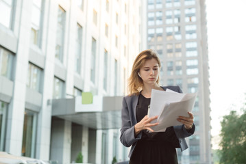 Concentrated stylish businesswoman standing on city street with documents reading them. Successful female manager dressed in elegant suit checking accounting on paper during coffee break outdoors.
