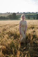 young pretty girl in yellow dress stands at field of ears in rays of rising sun. grove and village in background