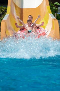 Caucasian boy and mom gliding down slide in waterpark. They enjoy the fun and holding hands wide open.