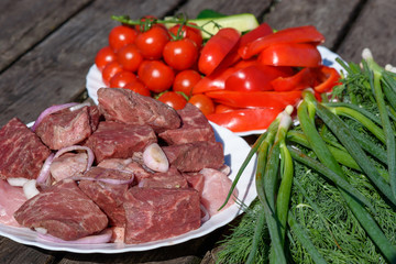 Cooking on fresh air. The plates with fresh meat, tomatoes and greenery are standing on the wooden table in the countryside getting ready for preparing.