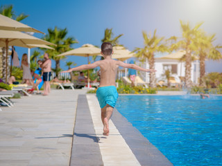 Caucasian boy having fun in swimming pool at resort. He is running along swimming pool. His arms are wide open.