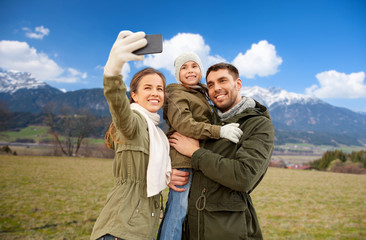 family, leisure and people concept - happy mother, father and little daughter taking selfie by smartphone over alps mountains in autumn background