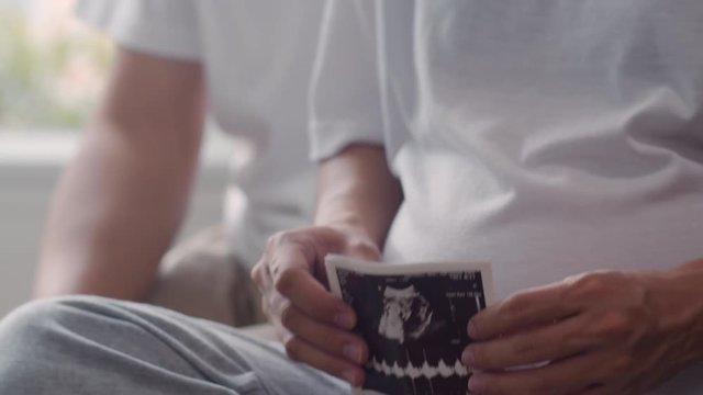 Young Asian Pregnant couple show and looking ultrasound photo baby in belly. Mom and Dad feeling happy smiling peaceful while take care child lying on sofa in living room at home concept. Slow motion.