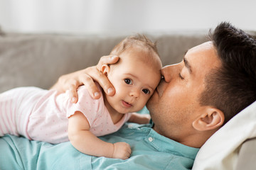 family, parenthood and fatherhood concept - middle aged father kissing little baby daughter lying on sofa at home - Powered by Adobe