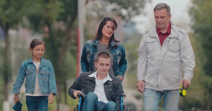 Happy parents walk with children. Son in a wheelchair for a walk with mom, dad, sister and dog. Portrait view.