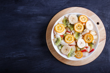 Vegetarian salad of bananas, apples, pears, kumquats and kiwi on black wooden background.
