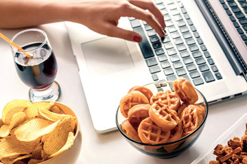 Girl works at a computer and eats fast food. Unhealthy food: chips, crackers, candy, waffles, cola. Junk food, concept.