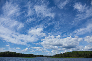 Background with thin white clouds in the blue summer sky over the lake. Forest on the far bank. With copy space