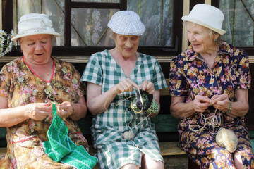 Three eldery seniors sit on a bench by the village house and knit. Sunny summer evening.