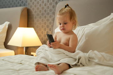 Cute toddler girl sitting on the bed and holding TV remote control	