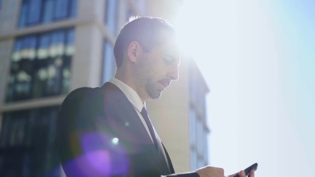Panning slow motion shot of middle aged businessman with stubble in black formal suit standing outdoors and text messaging on cell phone. Side view of man looking up thoughtfully in sunlight