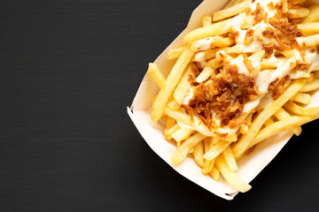 French fries with cheese sauce and onion in a paper box on a black background, overhead view. Flat lay, from above, top view. Copy space.