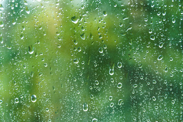 Close up of fresh water drops on window glass on background of green leaves. Raindrops on windowpane in summer day. Droplets on glass with blurry summer garden background. - Powered by Adobe