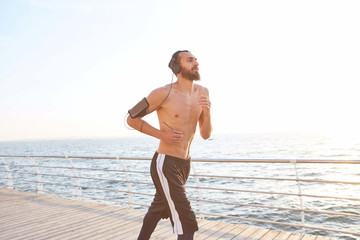 Fototapeta na wymiar Portrait of young handsome sporty guy with beard, running at the seaside, listen favorite mix on headphones. Enjoy the morning and jugging.