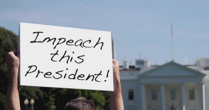 A man holds an IMPEACH THIS PRESIDENT protest sign in front of the White House on a sunny summer day.  	