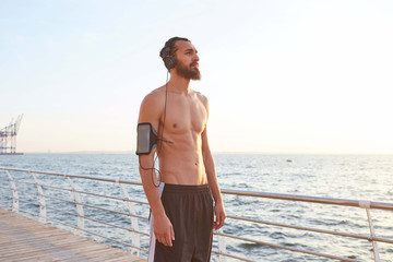 Fototapeta na wymiar Calming young attractive bearded guy has extreme sport at the seaside, rest after jogging, looking at the sea and listening songs on headphones, leads healthy active lifestyle. Fitness male model.