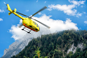 Fototapeta na wymiar Yellow helicopter on background of high Alps mountains wth green forest under blue cloudy sky.