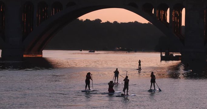 Washington, D.C. tourists explore the Potomac River using stand up paddle boards and kayaks on a hot summer day at sunset. The famous key bridge, a landmark, is in the background.