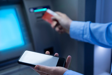 Man at ATM holding smartphone and credit cards, transferring funds / mobile upload. Night time 