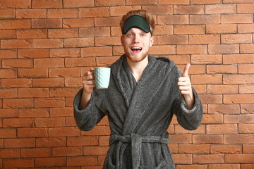 Happy young man with sleep mask and cup of coffee showing thumb-up gesture on brick background