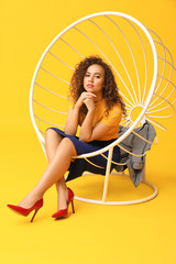 Stylish African-American woman sitting in armchair on color background