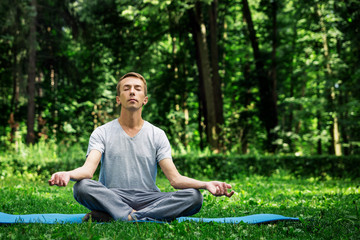 Young attractive man in sport clothes is meditating in the lotus position with a pacified face in the park against the background of green grass and trees.