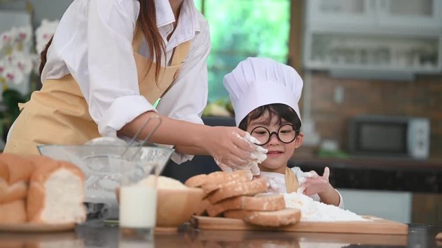 Cute little Asian boy with eyeglasses, chef hat and apron enjoy painting his mother face with dough flour bakery in home kitchen funny. Homemade food. Education and learning concept. Emotion portrait
