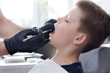 Ręce dentysty w jednorazowych czarnych gumowych rękawiczkach trzymają ekskawator dentystyczny. Chłopiec w wieku szkolnym siedzi na fotelu z otwartymi ustami. Lekarz ogląda zęby chłopca.