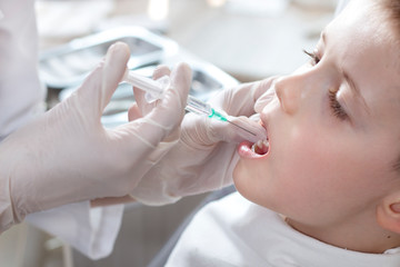 Chłopiec w gabinecie stomatologicznym podczas podawania znieczulenia. Dłonie stomatologa w białych rękawiczkach trzymają strzykawkę z igłą przy dziąśle małego pacjenta. - obrazy, fototapety, plakaty