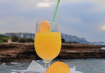 Close-up of a glass of orange juice with orange slices and rocks on the coast of the sea background