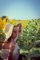 Portrait of young woman in white dress standing in the crops field of sunflowers in a sunny summer day wearing hat