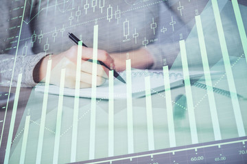 Hands writing information about stock market in notepad. Forex chart holograms in front. Concept of research. Multi exposure