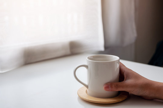 Closeup image of a hand holding a white cup of hot coffee on wooden table