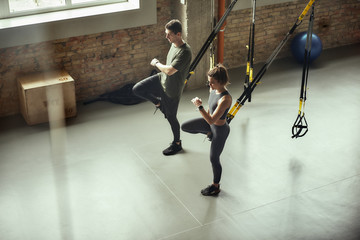 Exercising together. Young active couple in sportswear training legs with Trx fitness straps at gym.