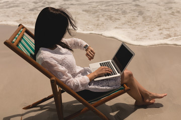 Young woman relaxing on sun lounger and looking smartwatch while using laptop 