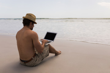 Young man with hat using laptop on the beach