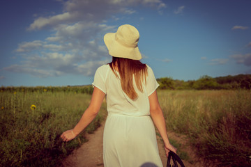 Portrait of young woman in white dress with hat standing in the dirt road in summer day walking away back view